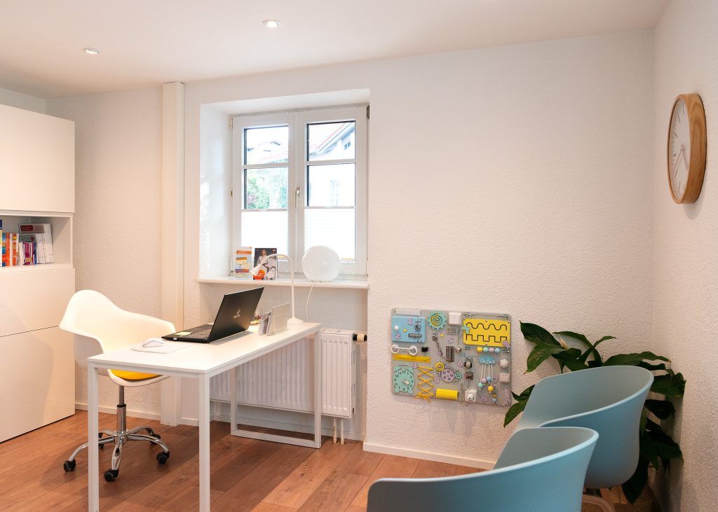 Sprechzimmer mit Arbeitsplatz, Patientenstühlen und einer Spielecke im Hintergrund