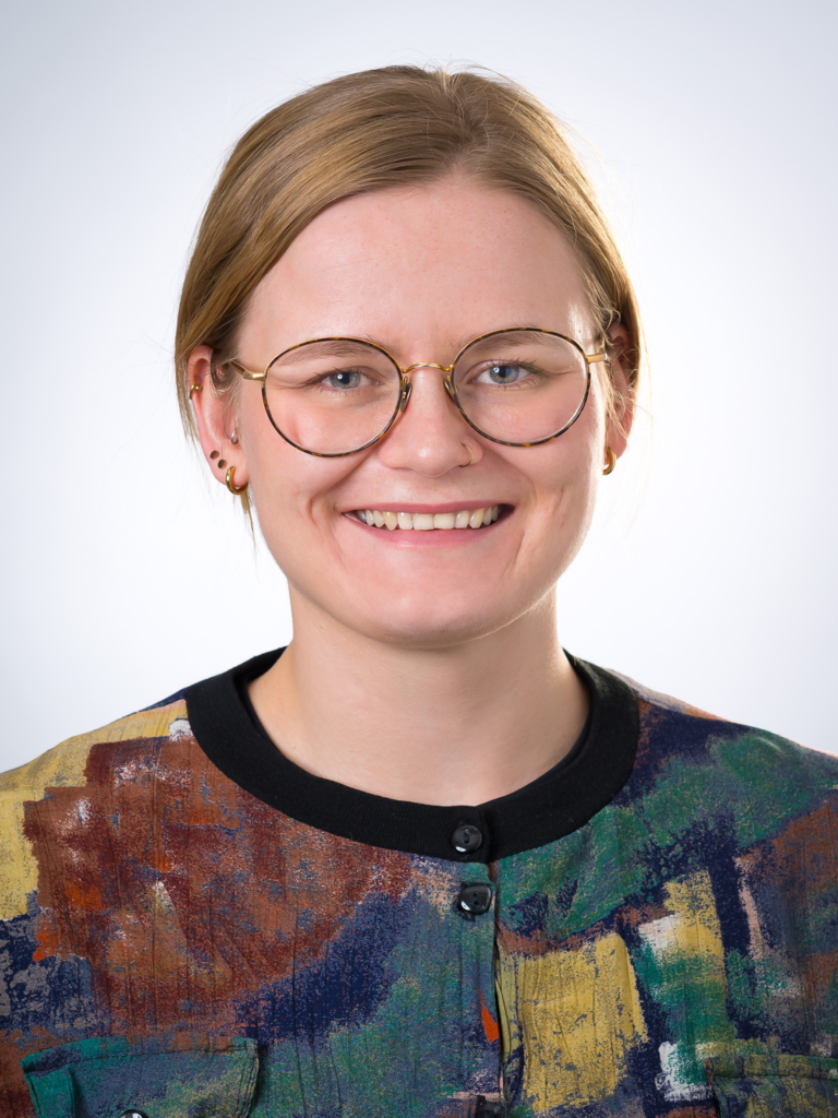 Porträt von Isabel Brand, lächelnd mit Brille und blonden Haaren in einem bunten Oberteil