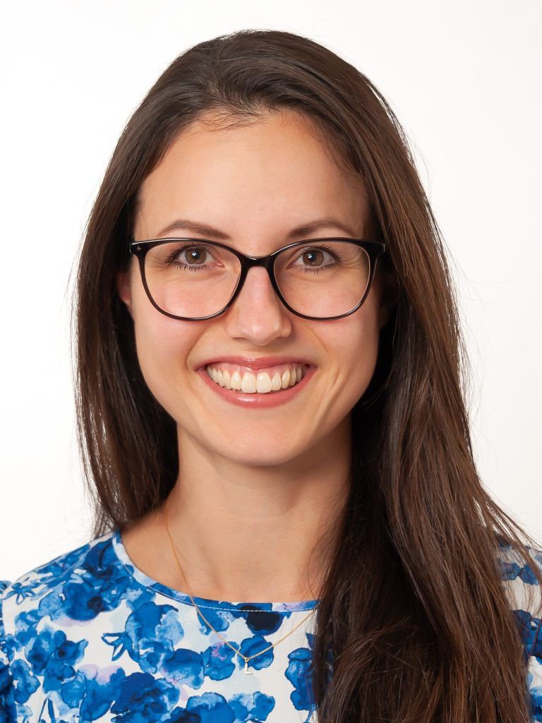 Porträt von Maria Betz, lächelnd mit Brille und langen braunen Haaren in einer blau geblümten Bluse