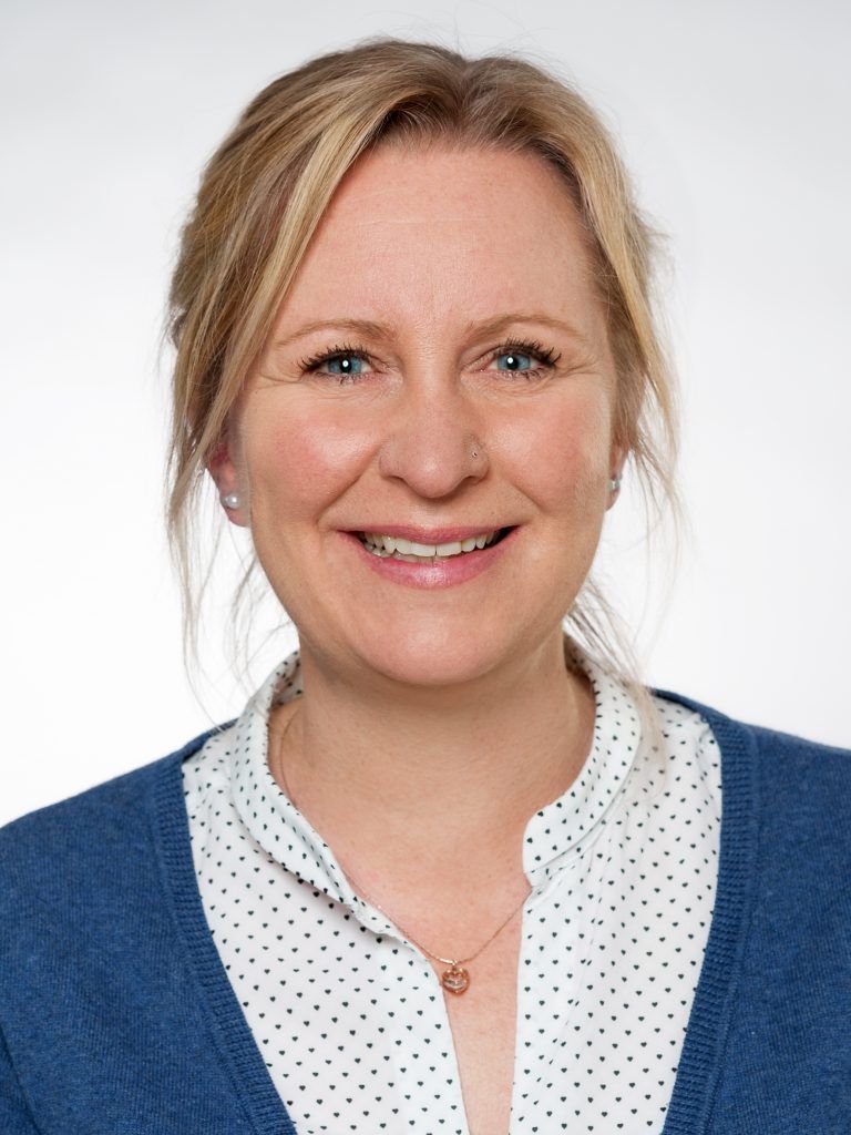 Porträt von Sandra Sterzik, lächelnd mit blonden Haaren in einer gepunkteten Bluse und blauer Strickjacke